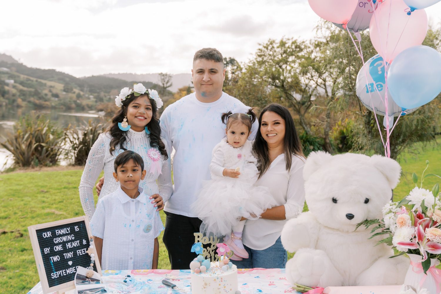 Family of five poses at a gender reveal party with pink and blue balloons, a white teddy bear, and a decorated table outdoors.