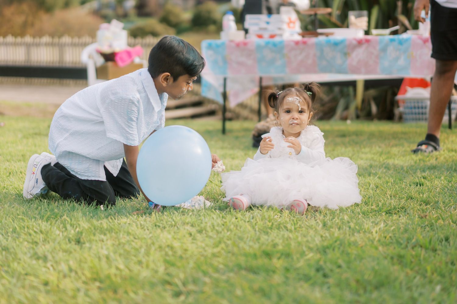A boy in white shirt plays with a blue balloon beside a toddler girl in a white tutu dress on green grass.