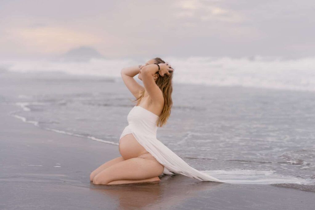 Pregnant woman in white dress kneeling on beach at sunset, waves behind her.