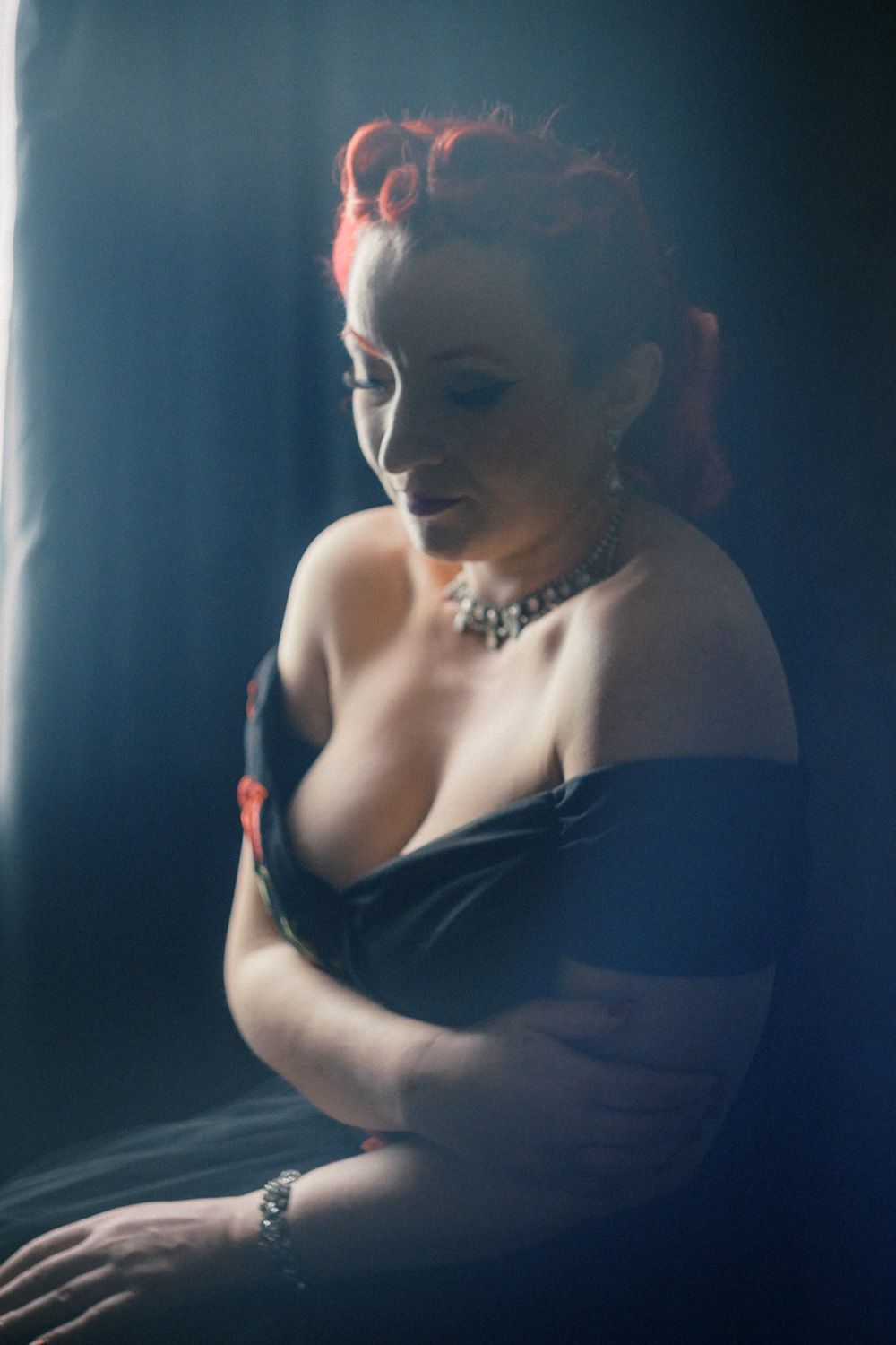 Dramatic portrait of a redhead woman in a black off-shoulder dress with a statement necklace, posed in moody blue lighting.