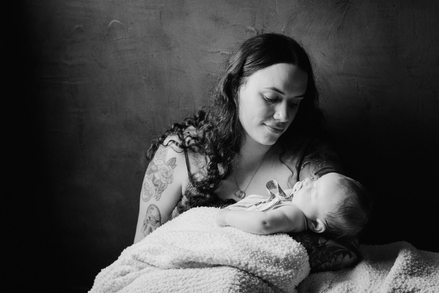Black and white photo of tattooed mother tenderly breastfeeding newborn baby wrapped in soft blanket.