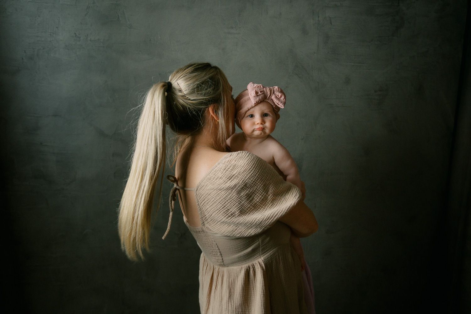 A blonde mother in a beige dress holds her baby girl wearing a pink bow against a dark studio backdrop.
