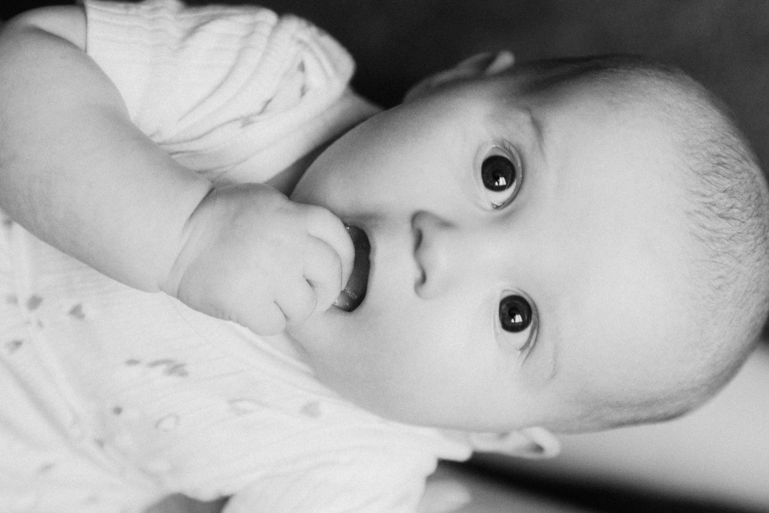 Black and white close-up portrait of a curious baby with bright eyes, gently touching their mouth.
