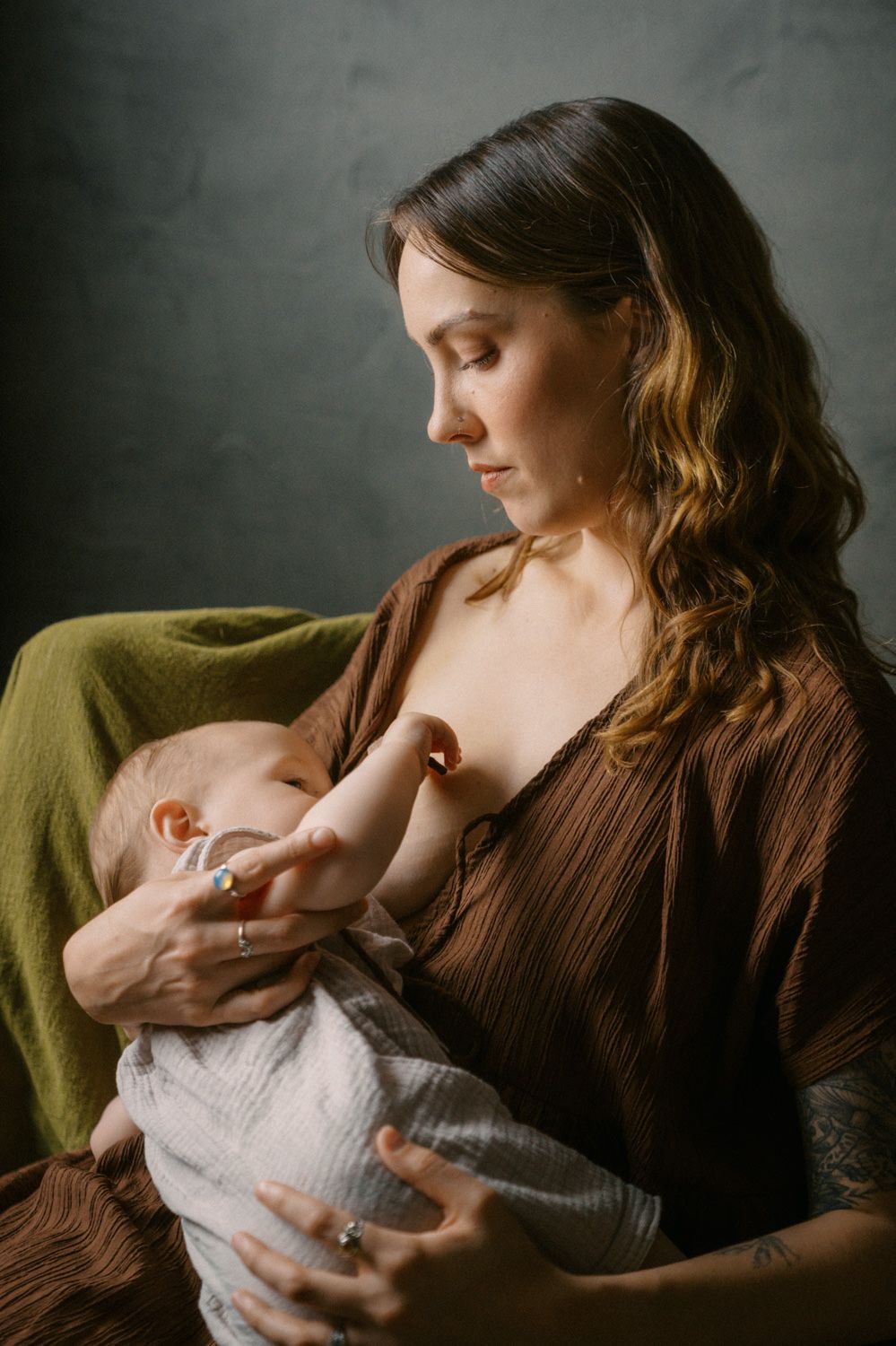 A mother tenderly breastfeeds her newborn baby while seated, wearing a brown wrap dress against a gray backdrop.