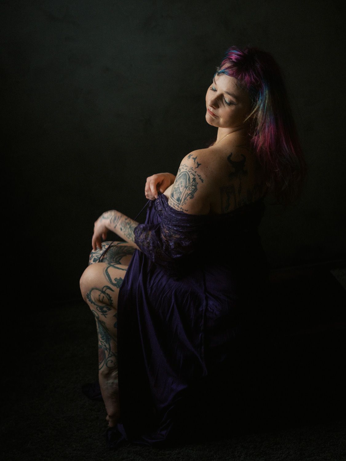 A tattooed woman with colorful purple-pink hair poses in a purple dress against a dark background.
