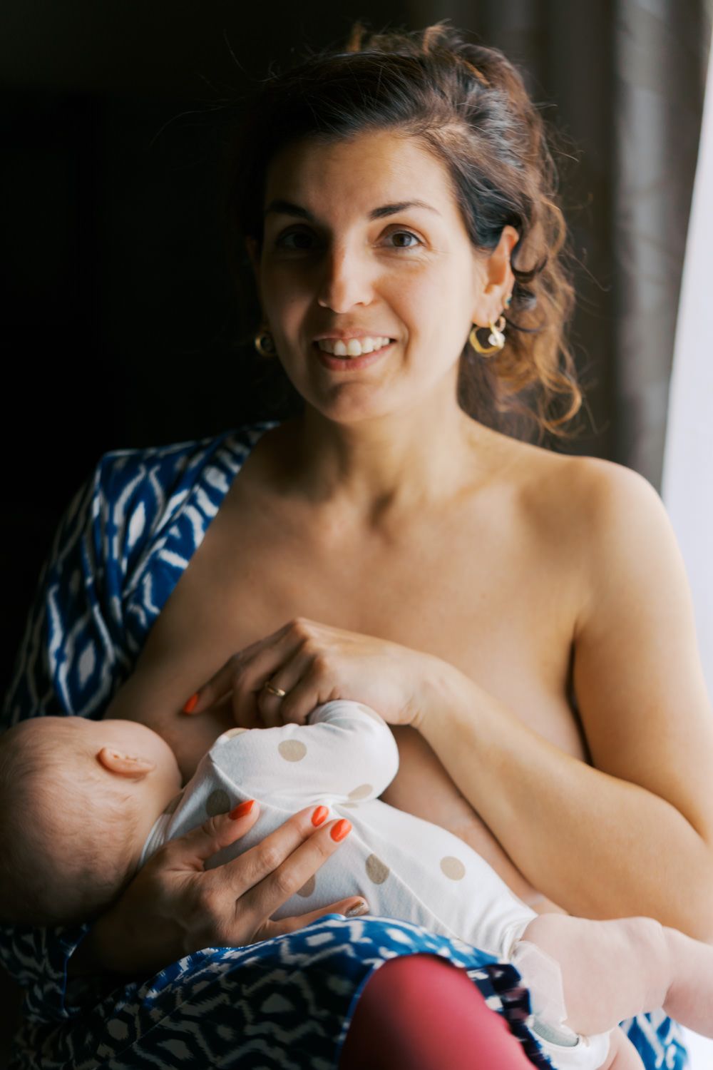 Smiling mother breastfeeding her newborn baby while sitting in a blue patterned chair near a window.