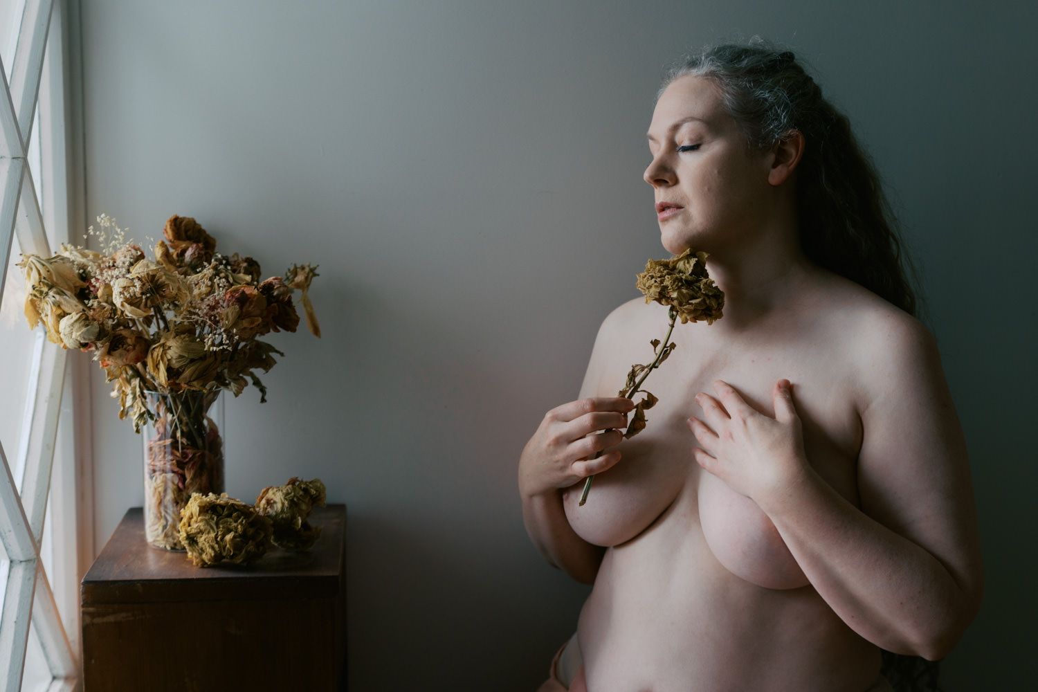 A topless woman with gray hair holds dried flowers near a window beside a vase of dried botanicals.