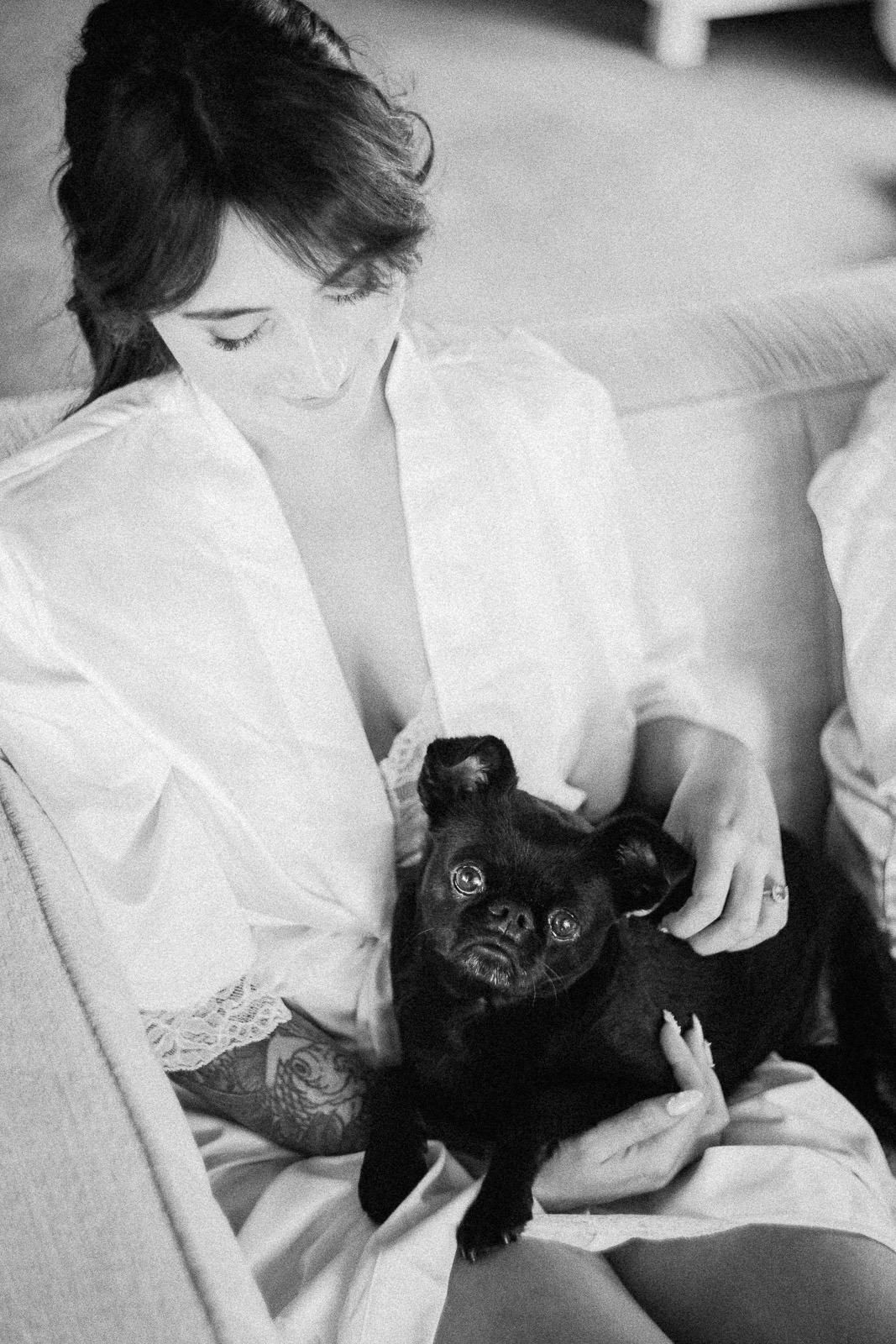 Black and white photo of a woman in a white robe sitting on a sofa, gently holding a small black pug dog.