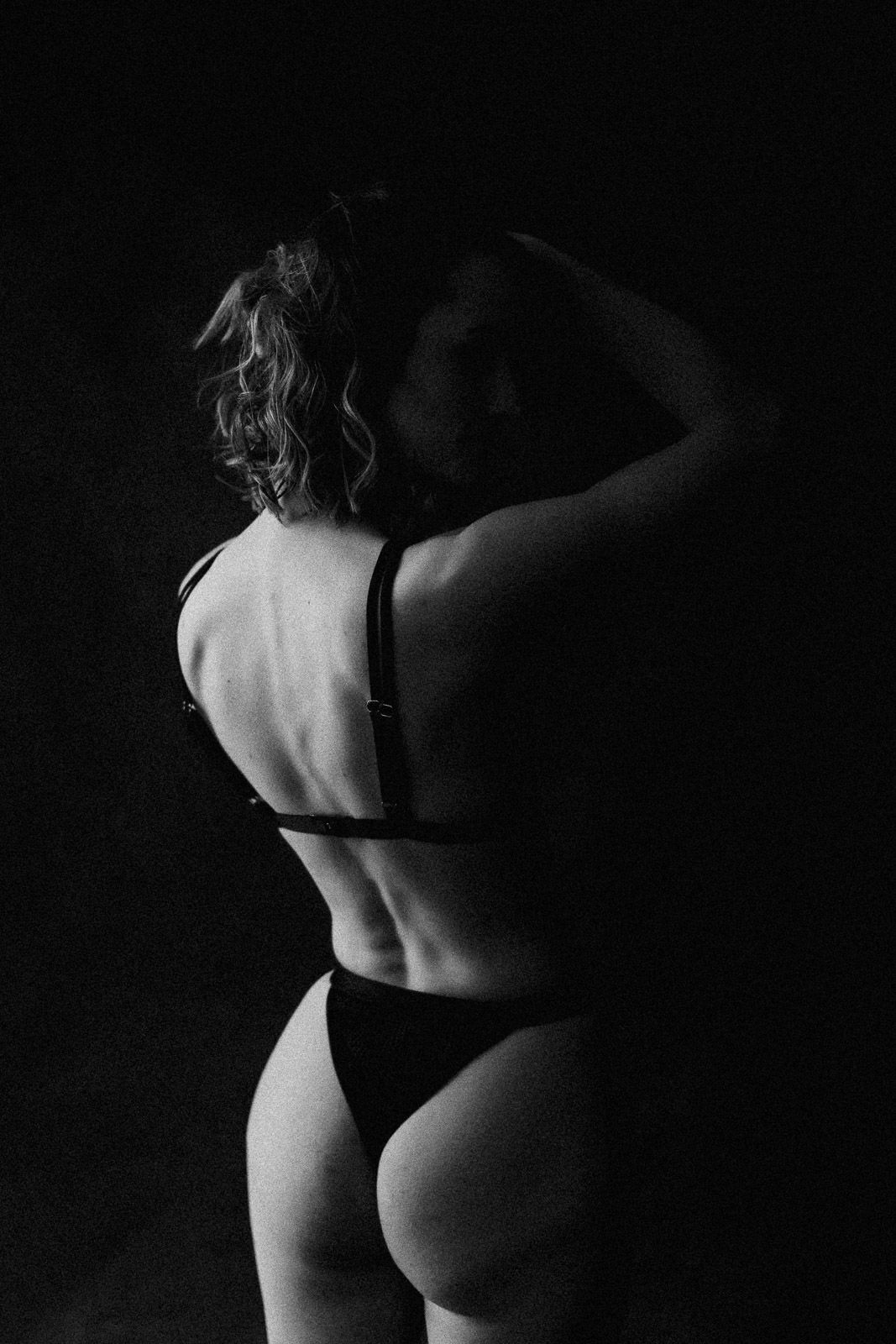 Black and white artistic photo of a woman in lingerie posing with back to camera against dark background.