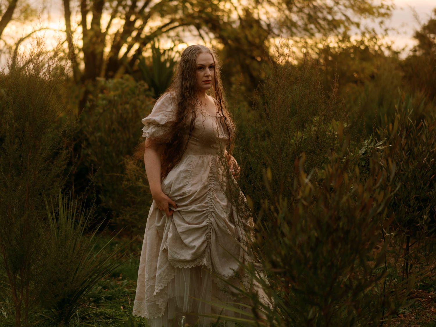 Woman in antique white gown standing among lush green bushes at golden hour.