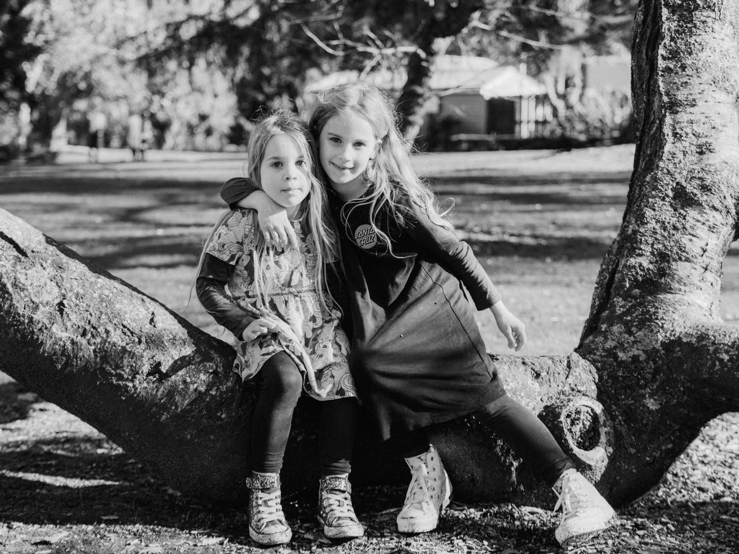Black and white photo of two young blonde girls sitting together on a large tree branch.