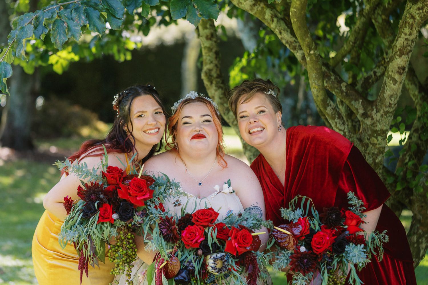 Three women in red and yellow velvet dresses smile holding lush red rose bouquets outdoors under green trees.