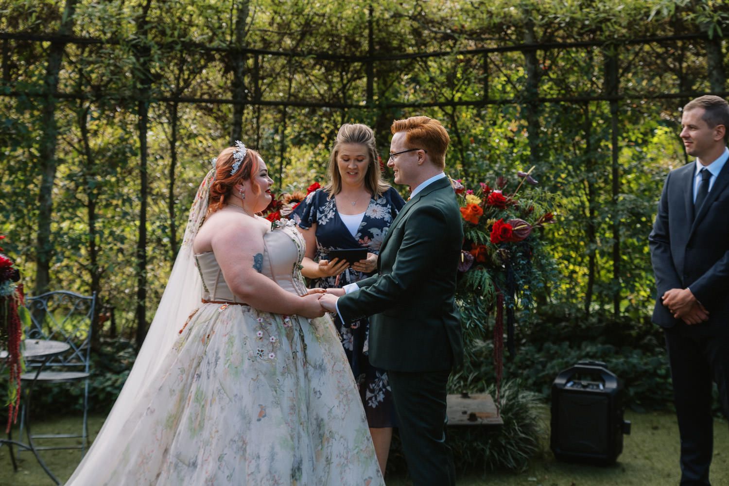 Couple exchanging vows at outdoor wedding ceremony with officiant and floral arch backdrop.
