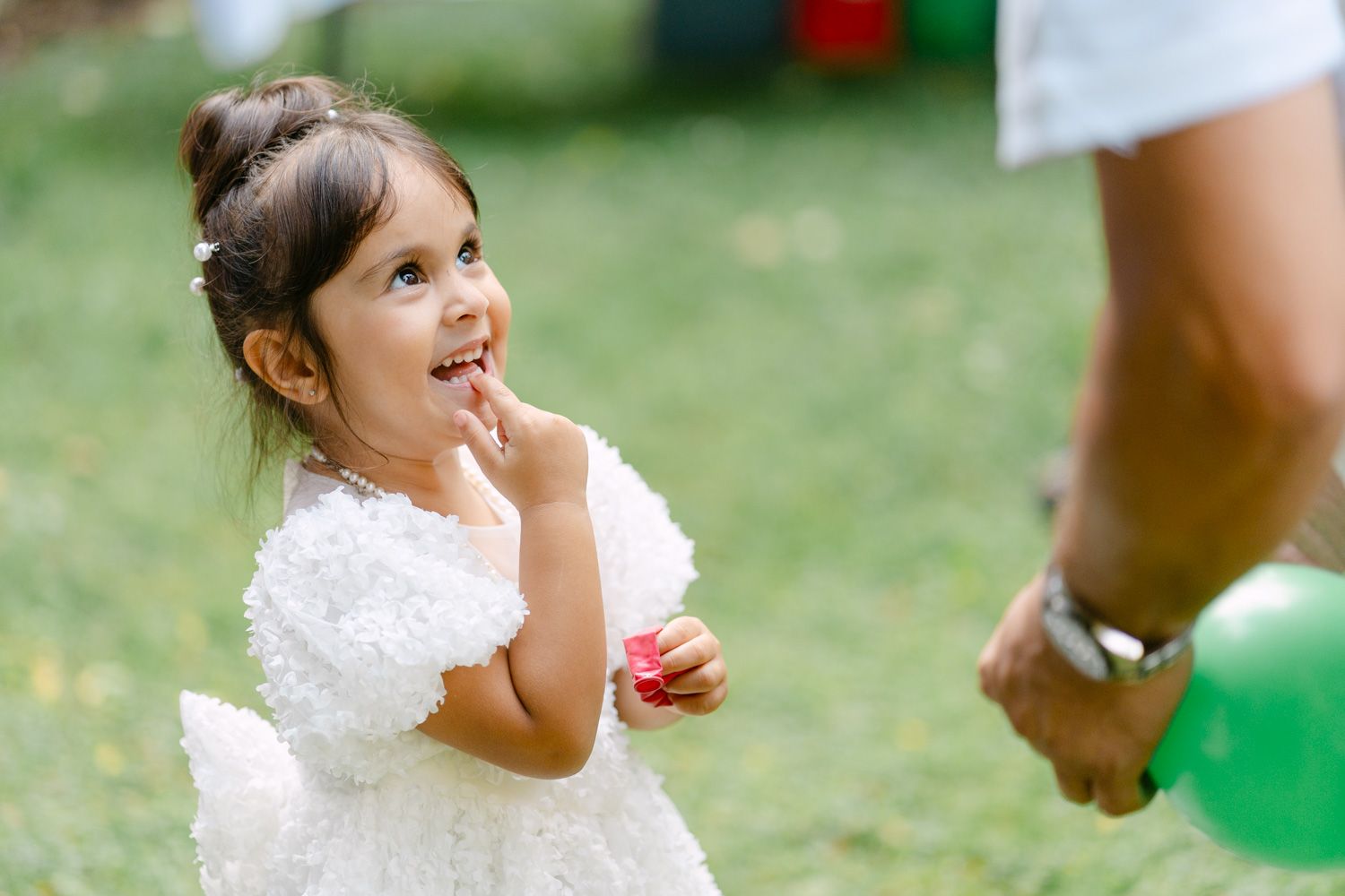 Adorable little girl in white dress smiling outdoors, holding a flower near green balloons at a garden party.