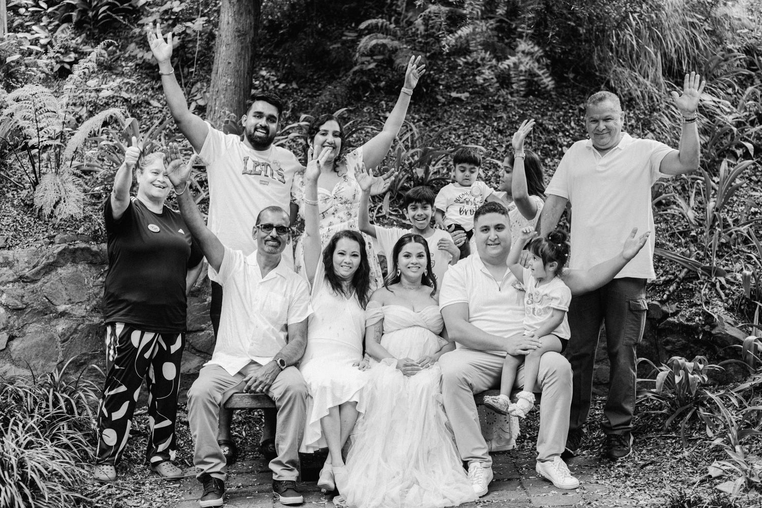 Black and white family photo outdoors with group waving and smiling, featuring a bride and pregnant woman seated.