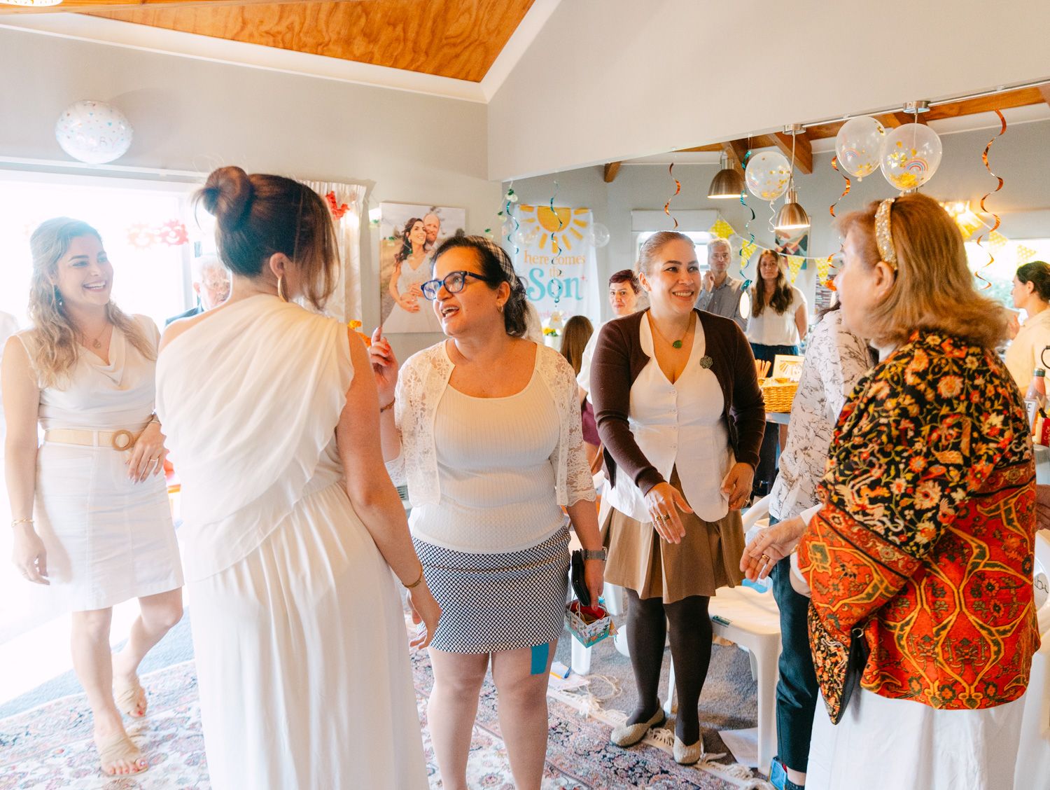 Women socializing at an indoor baby shower party with balloons and decorations in a bright, welcoming room.