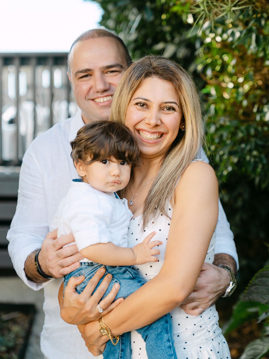 Happy family portrait of parents smiling and holding their young toddler outdoors among green foliage.