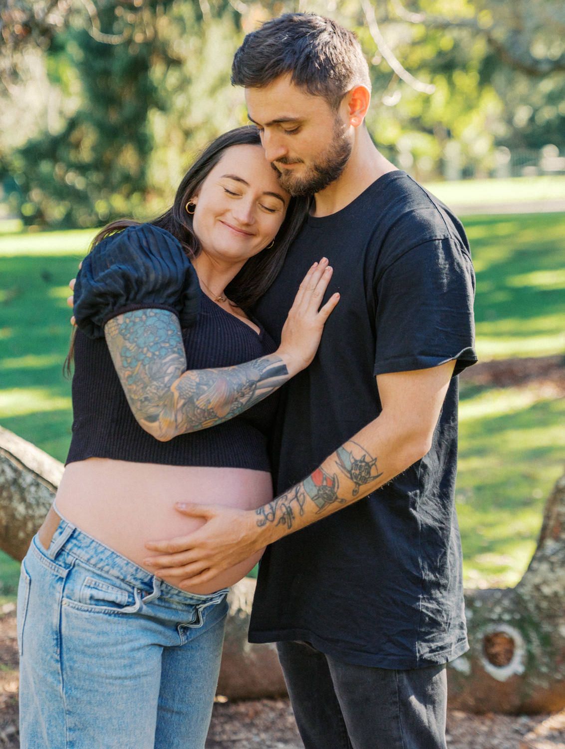 A tattooed couple shares a tender embrace outdoors, with the woman showing her pregnant belly in a sunny park setting.