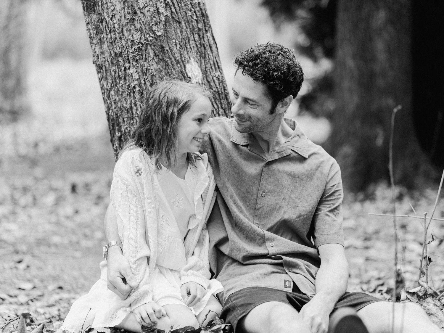 Black and white photo of a smiling couple sitting together against a tree in a forest setting.