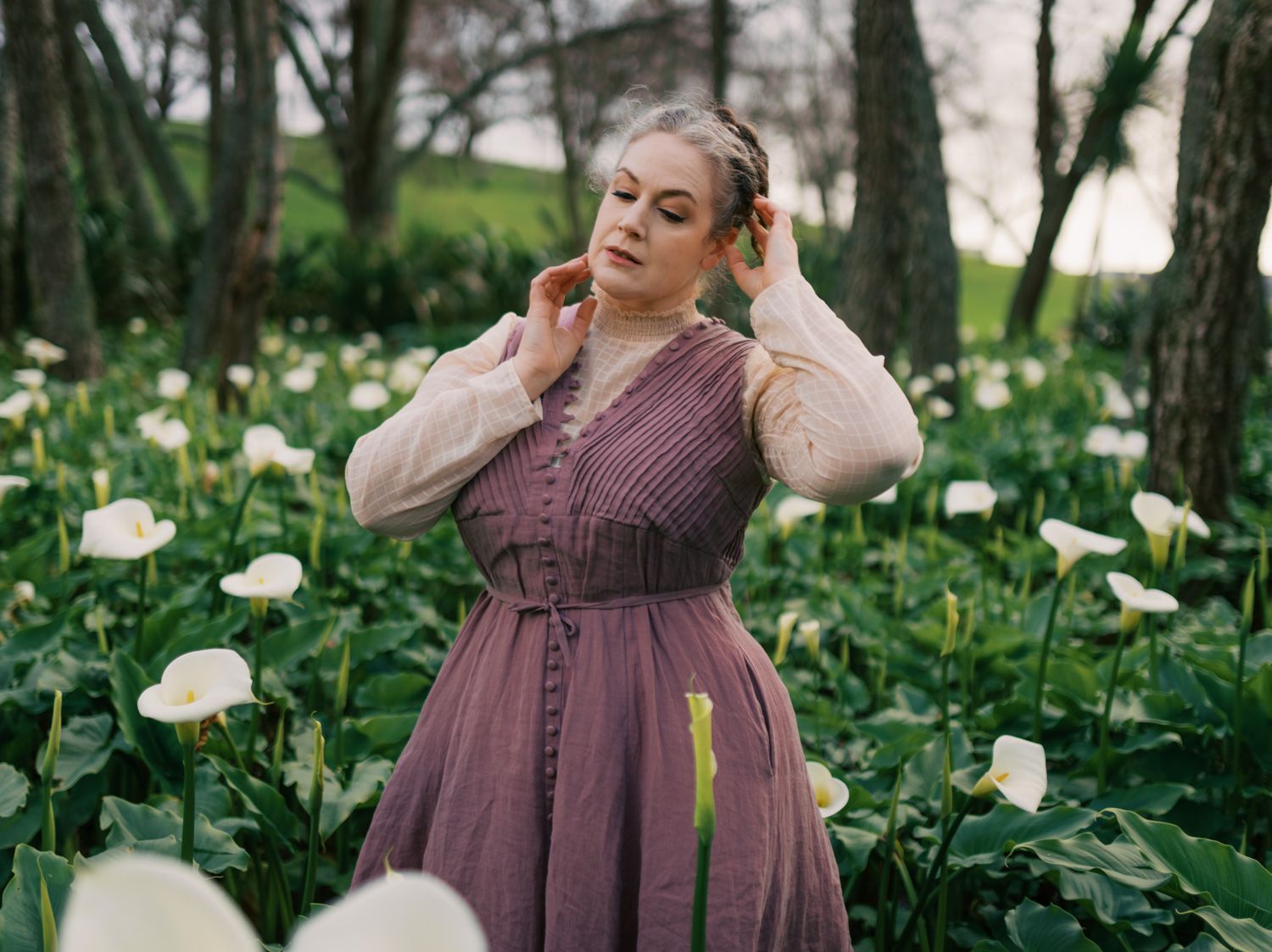 Woman in mauve dress among white calla lilies in lush green garden, hands in hair, serene outdoor portrait.