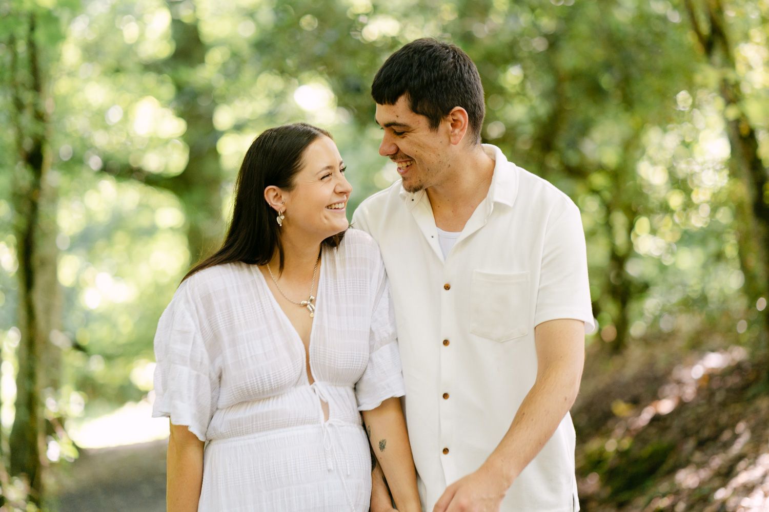 Smiling pregnant couple holding hands on a tree-lined path, wearing white outfits in a lush green forest setting.