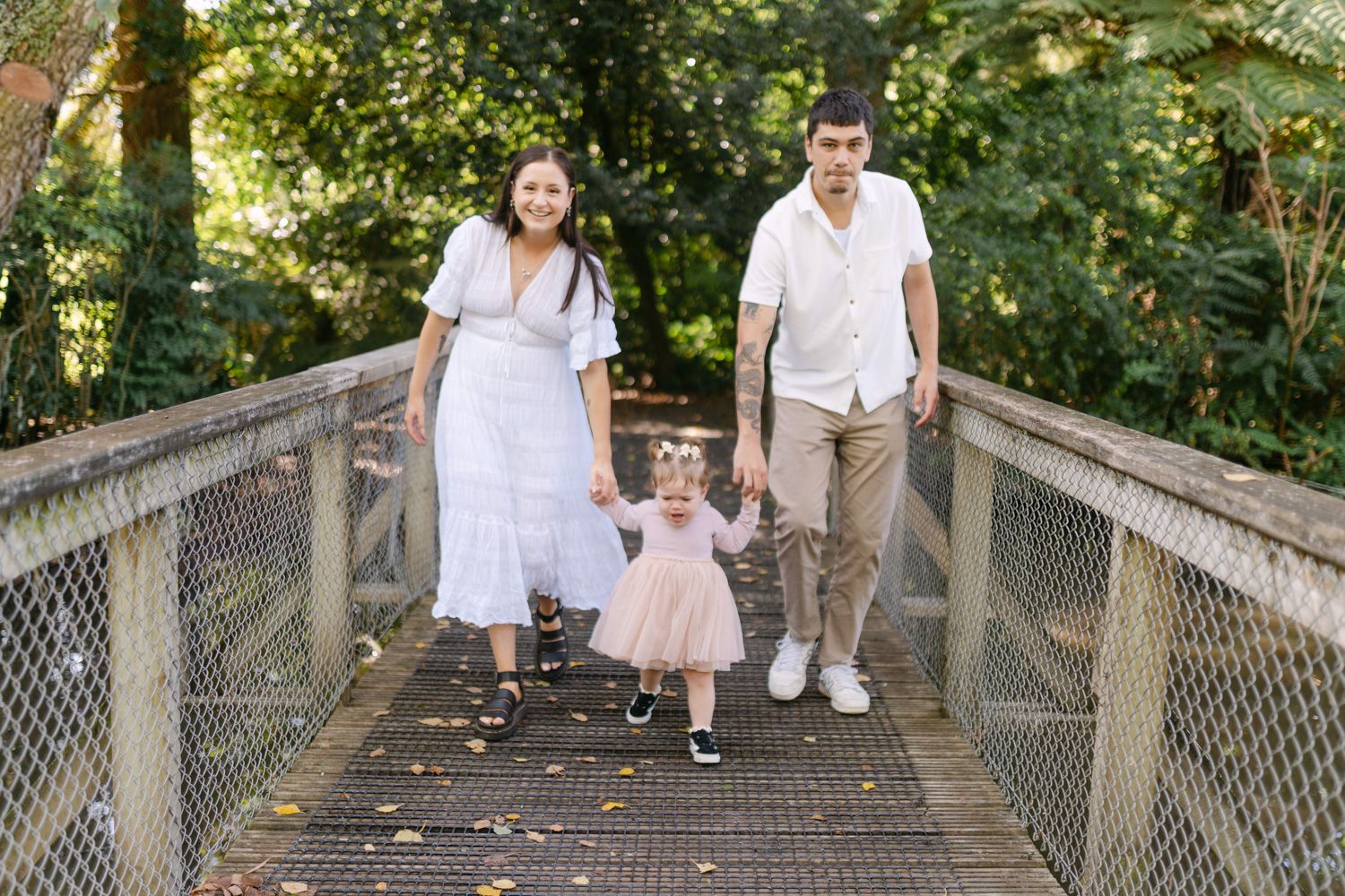 Family of three holding hands on a wooden bridge surrounded by lush green trees, parents in white outfits with toddler in pink dress.