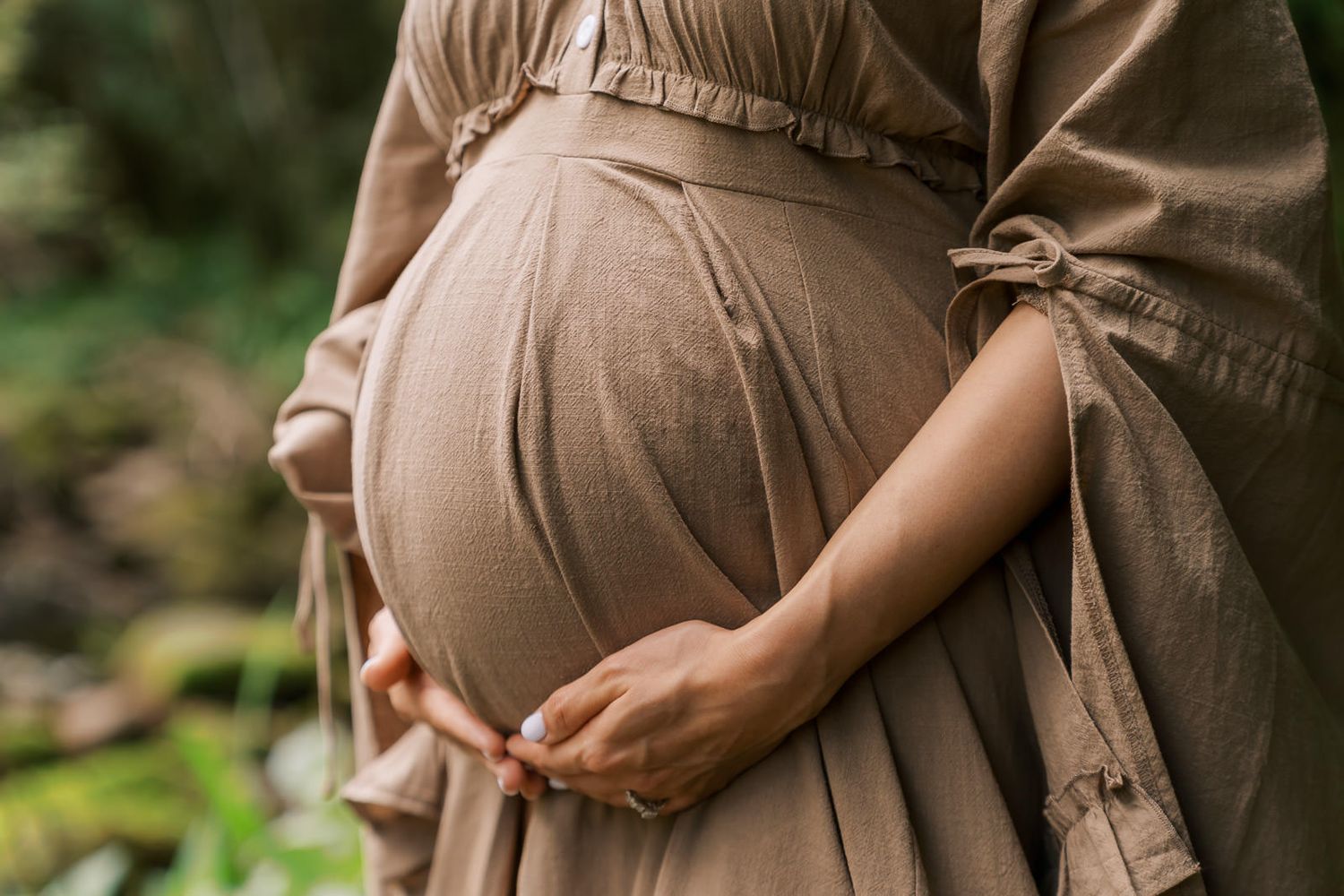 Pregnant woman in beige linen dress cradling baby bump outdoors in lush green nature setting.