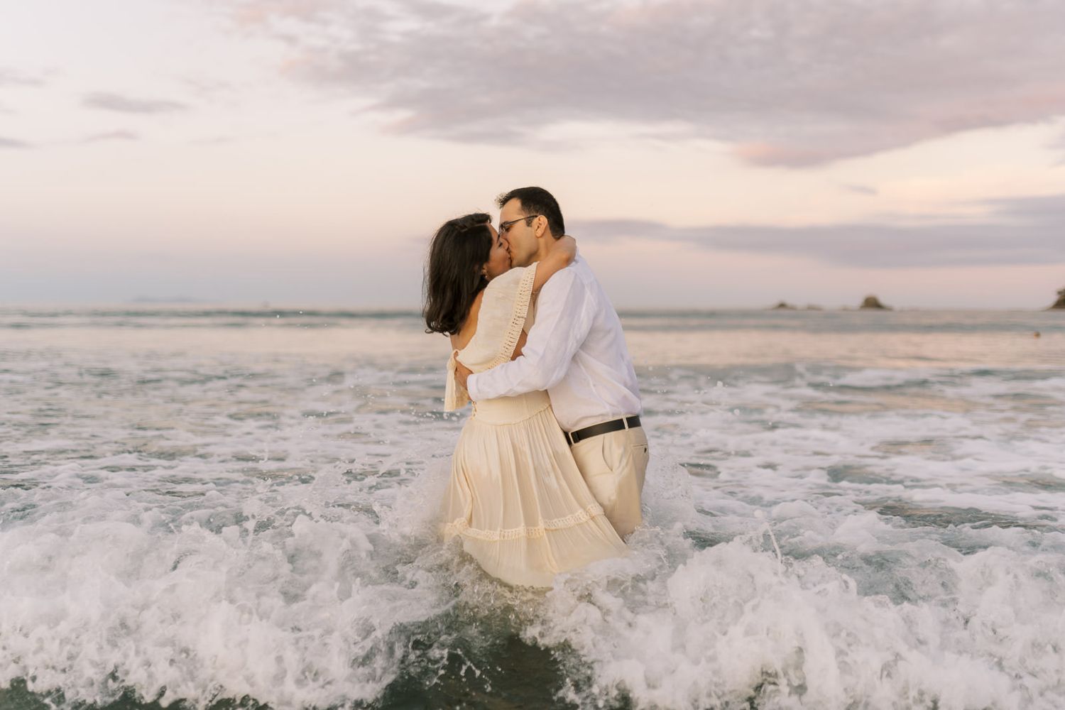 Couple sharing a romantic kiss in ocean waves at sunset, wearing white outfits on a beautiful beach.