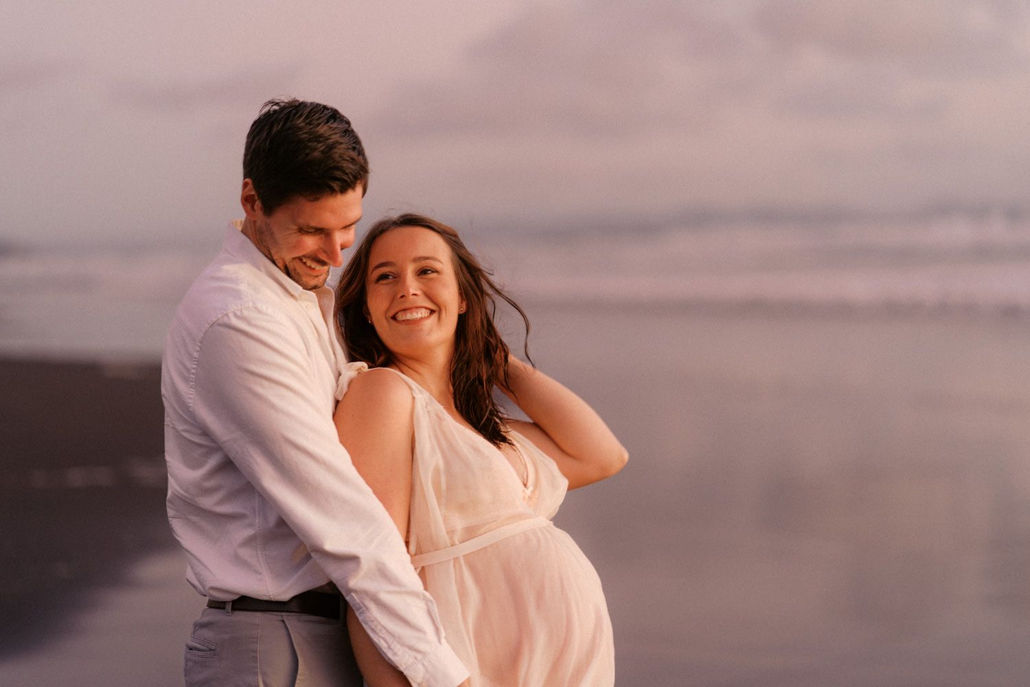 Couple embracing on a beach at sunset, woman in white dress smiling, waves in background.