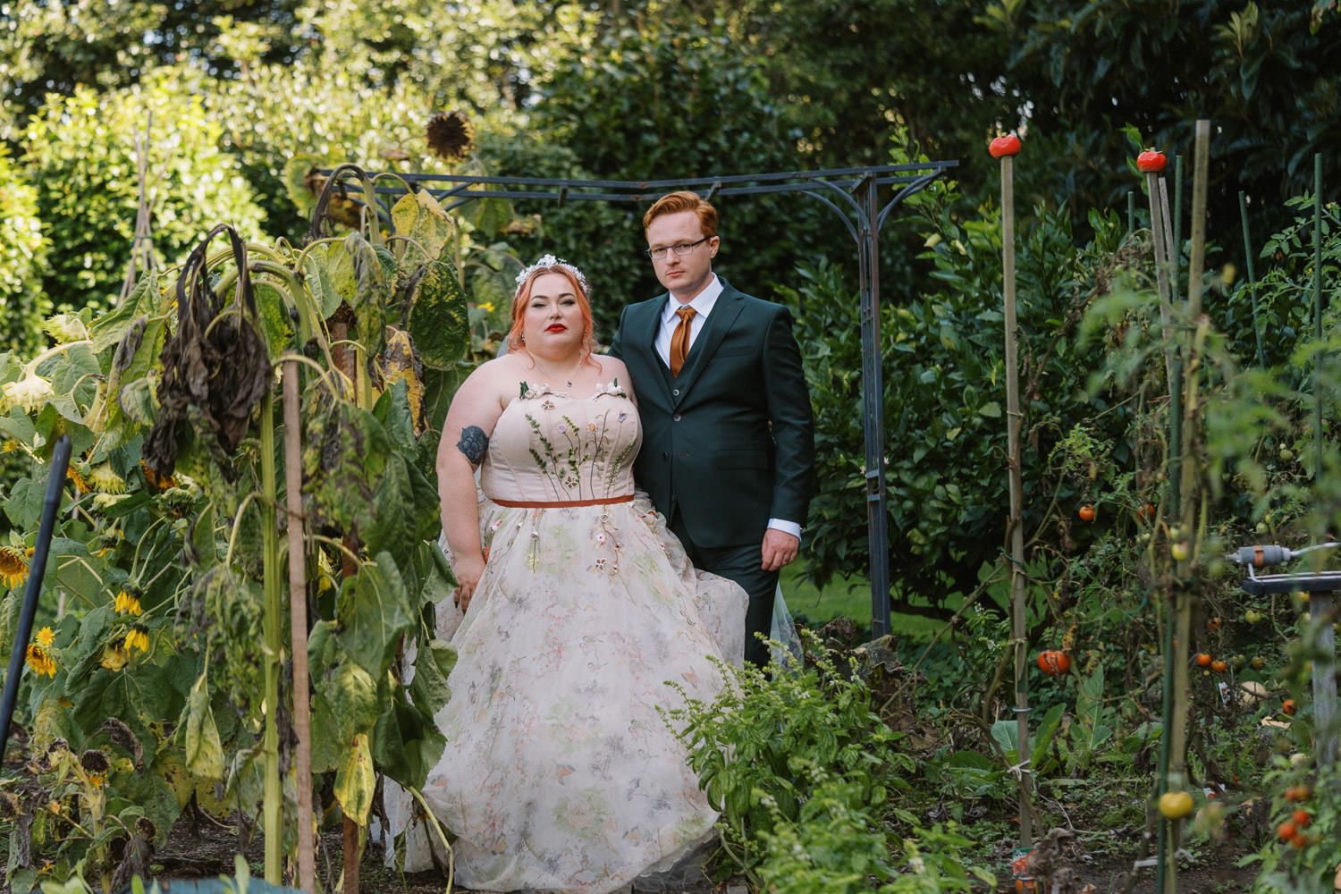 Bride and groom standing in a lush garden wedding setting, bride in floral gown, groom in dark green suit.