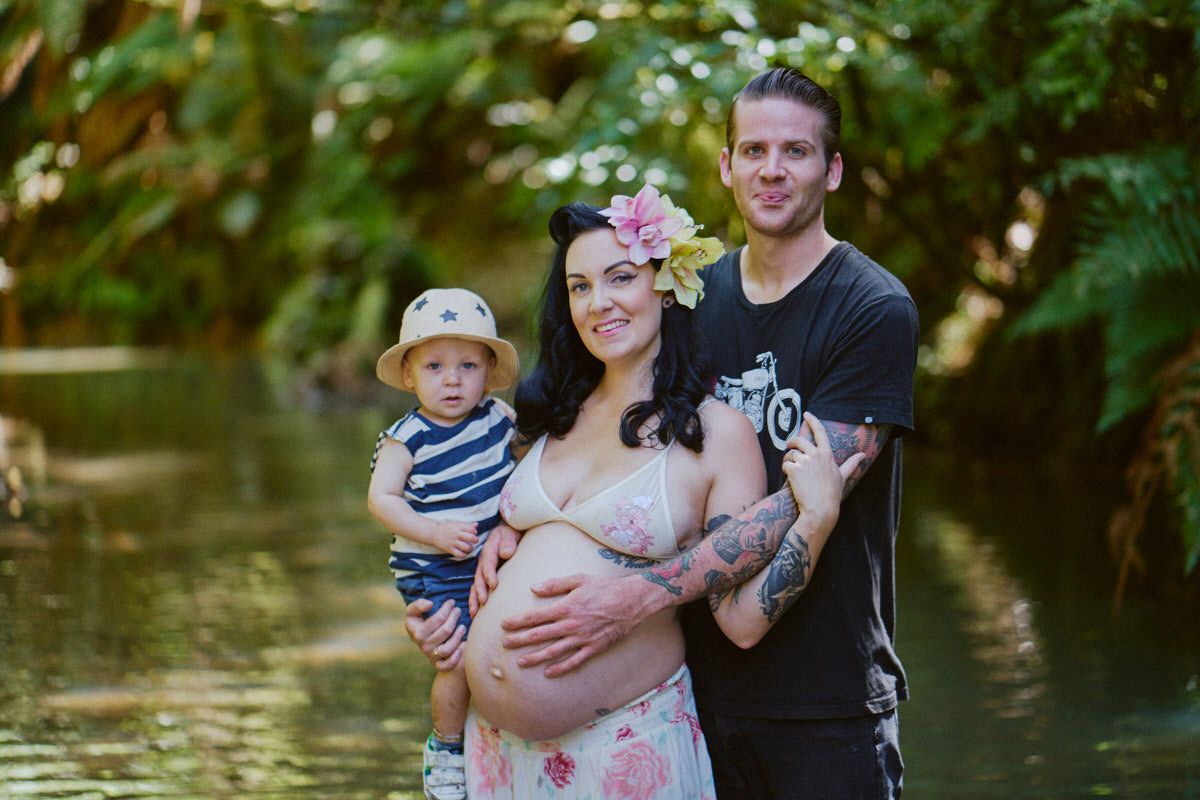 Pregnant woman with flower crown poses with tattooed partner and toddler in a lush green creek setting.