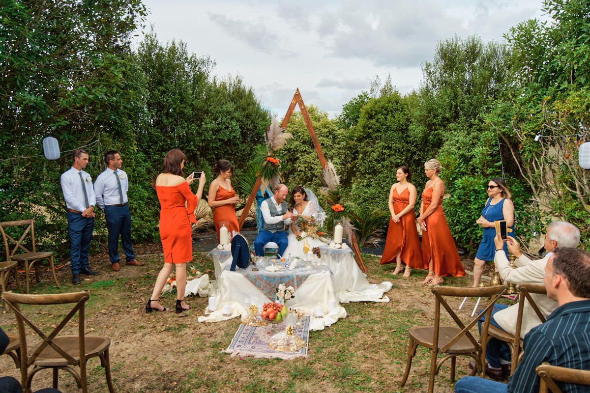 Outdoor wedding ceremony in a garden with guests in orange dresses around a decorated table under a triangular arch.