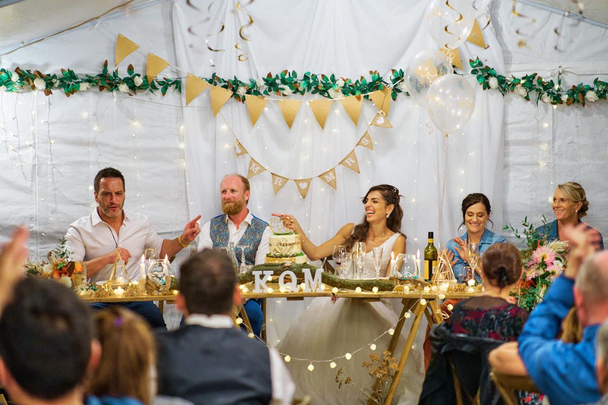 Wedding party laughing and toasting at elegantly decorated reception table under a white tent with bunting.