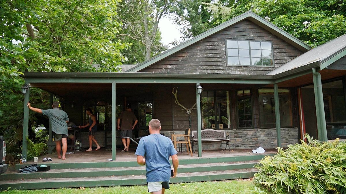 Two men approach a rustic wood-sided cottage with a covered porch surrounded by lush green trees.