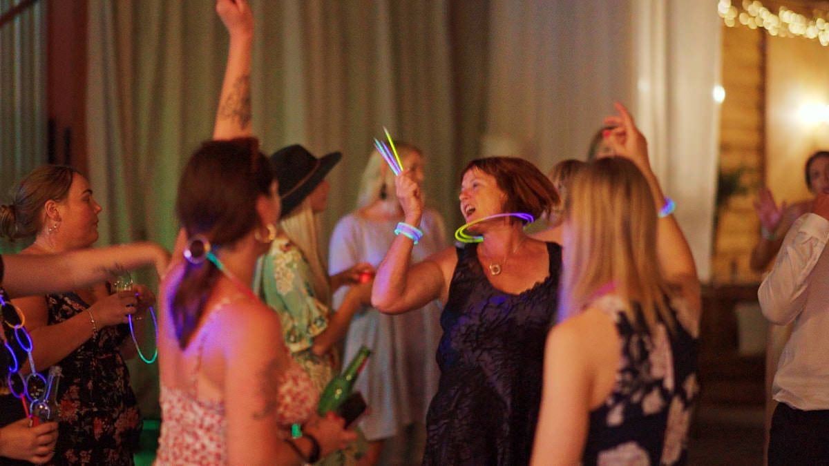 Women dancing at a party with glow sticks and neon accessories under warm festive lighting.
