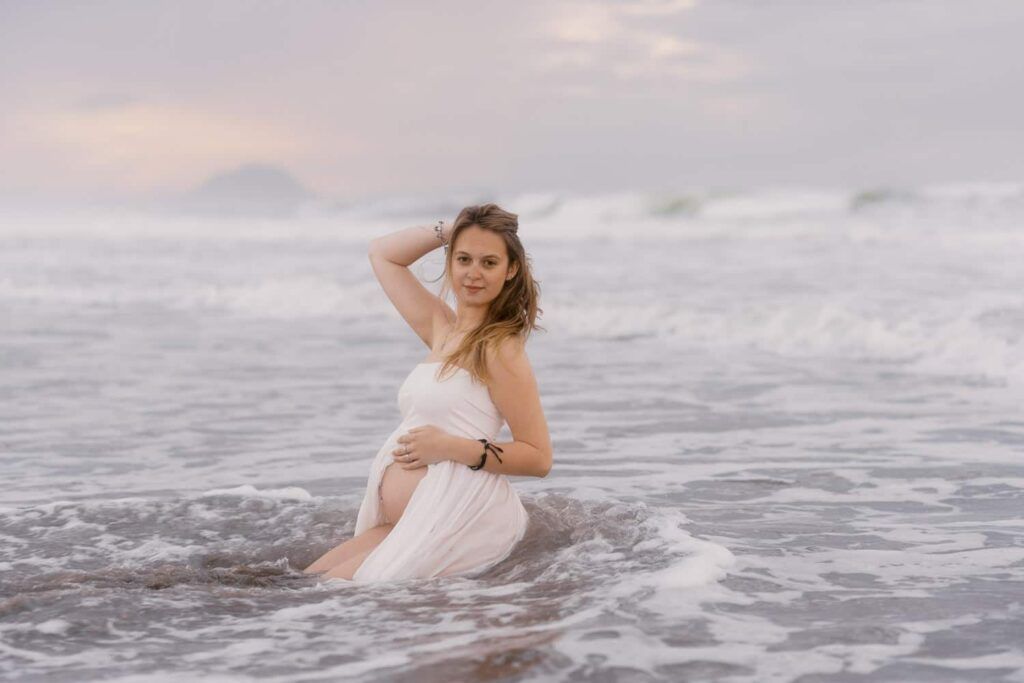 Pregnant woman in white dress posing in ocean surf during golden hour maternity photography session.