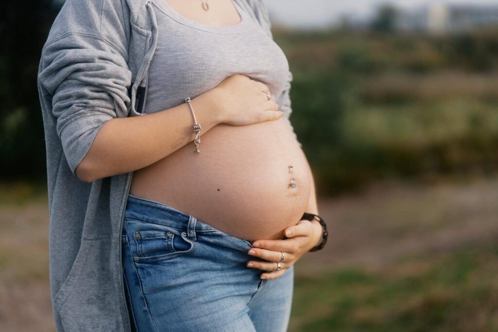 Pregnant woman in gray hoodie and jeans cradling her bare baby bump outdoors in a natural setting.