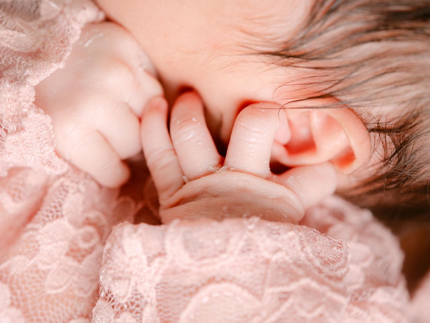 Close-up of a sleeping newborn baby in pink lace outfit, tiny hands near face, soft pink tones.