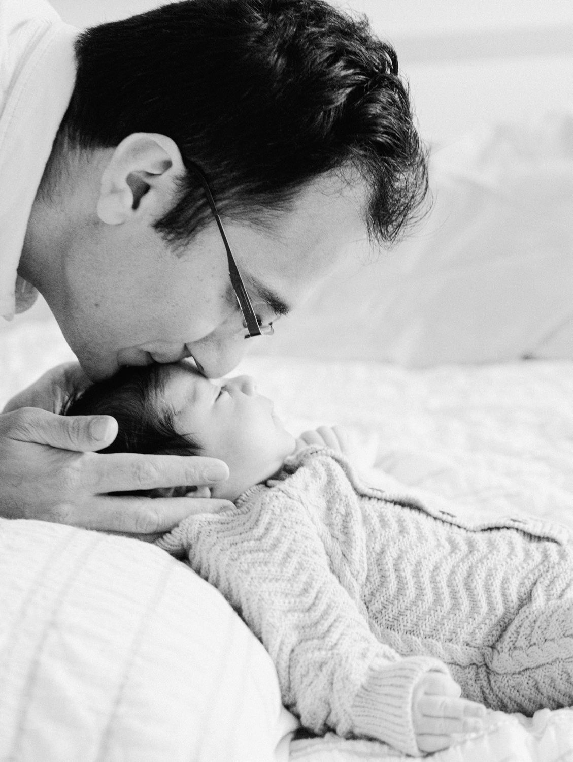 Black and white photo of a father lovingly kissing his newborn baby on a white bed.