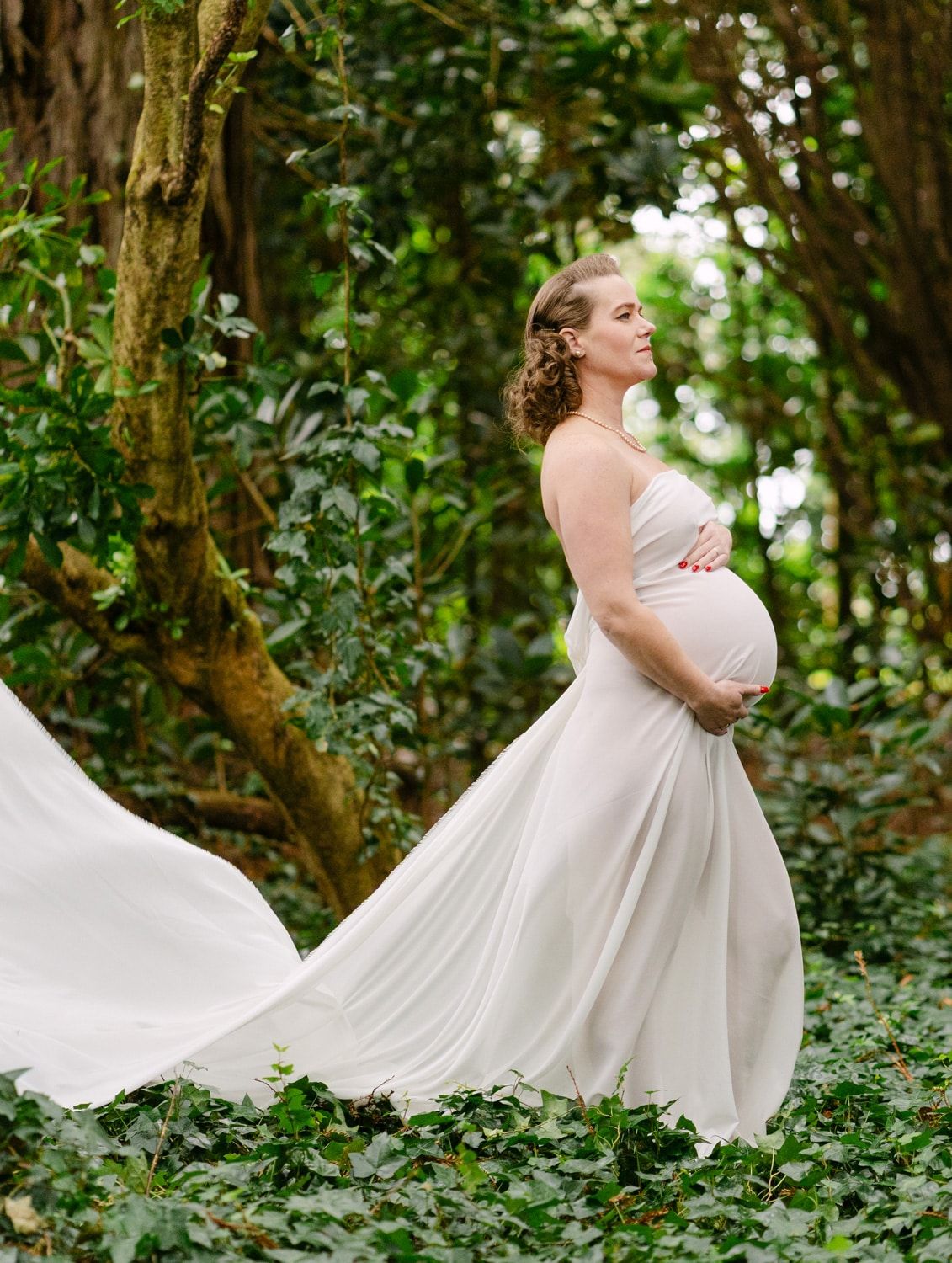 Pregnant woman in flowing white gown stands gracefully among lush green ivy and trees in an elegant outdoor maternity portrait.