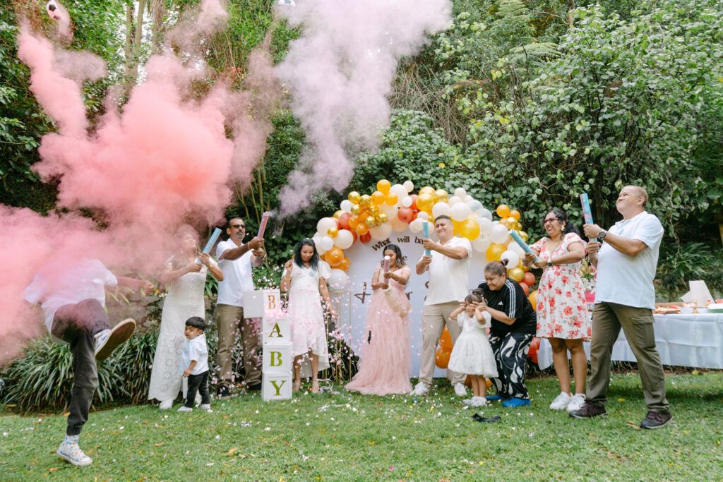Gender reveal party with pink smoke, balloons, and excited guests celebrating outdoors in a lush garden setting.