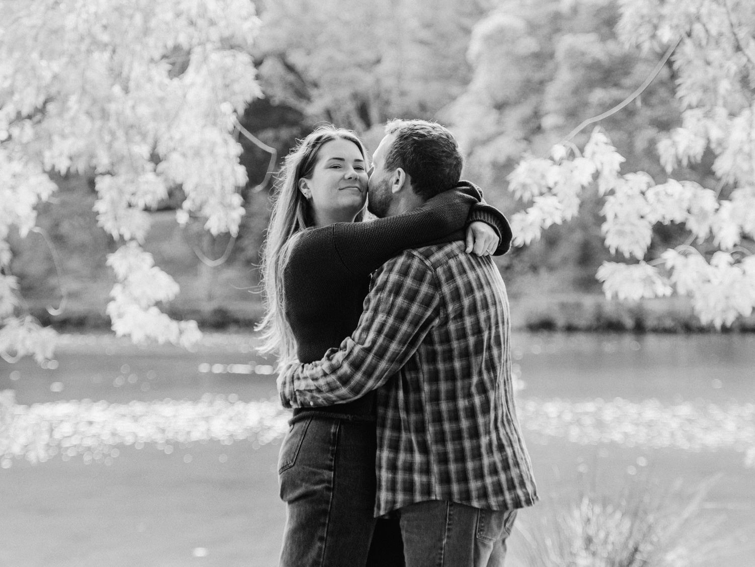 Black and white photo of a couple embracing and kissing near a lake with blooming trees in background.