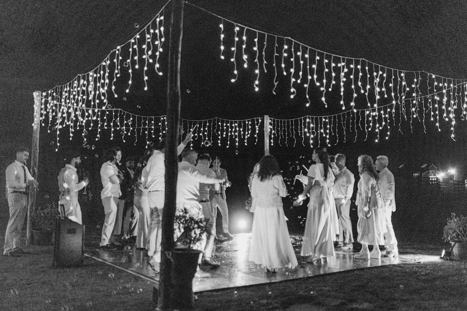 Black and white photo of elegant outdoor evening party under canopy with string lights and guests in white attire.