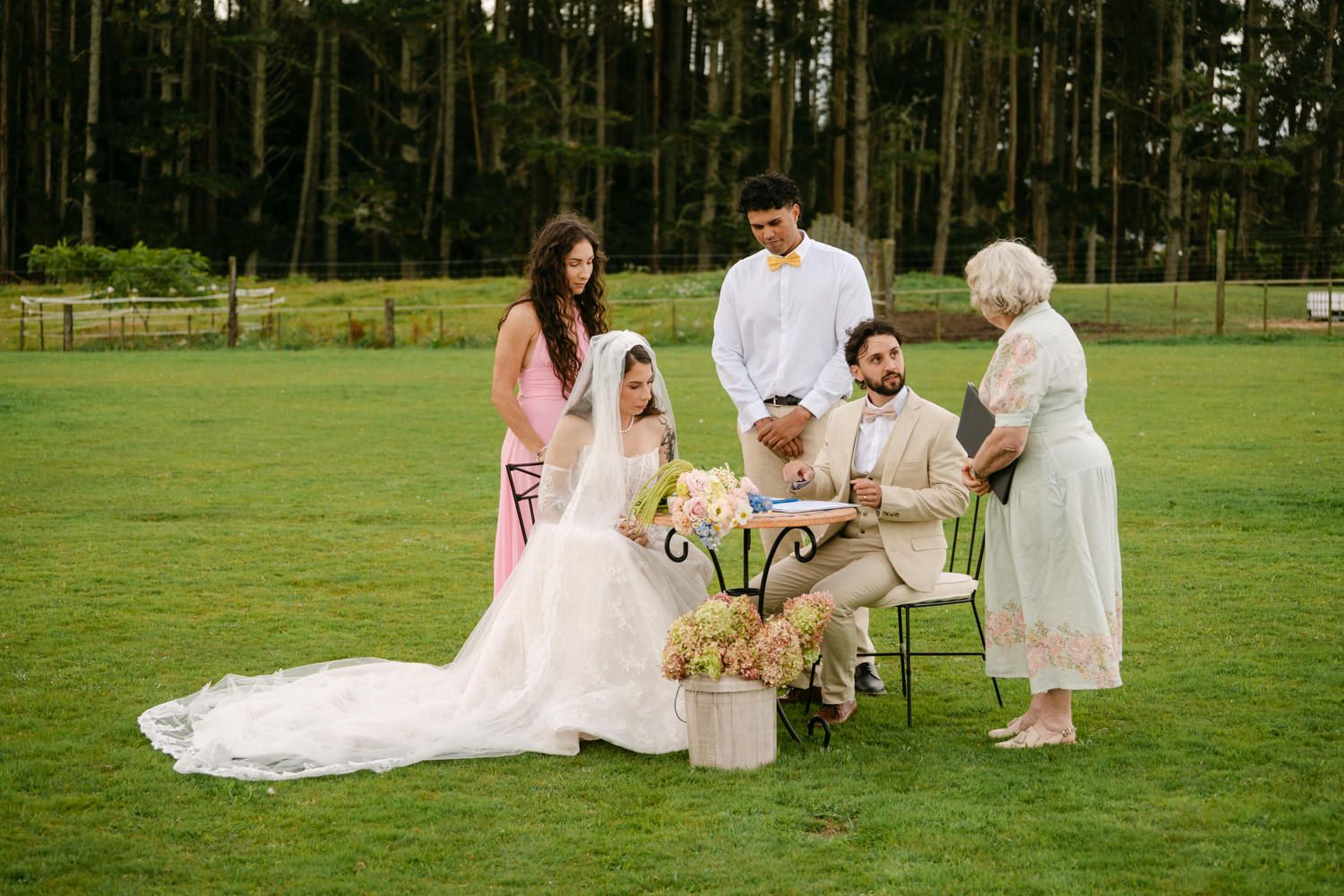 Outdoor wedding ceremony on green lawn with bride, groom, officiant, and guests signing documents near floral arrangements.
