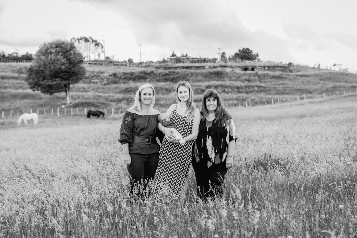 Three women standing together in a field with horses grazing in the background, black and white photo.