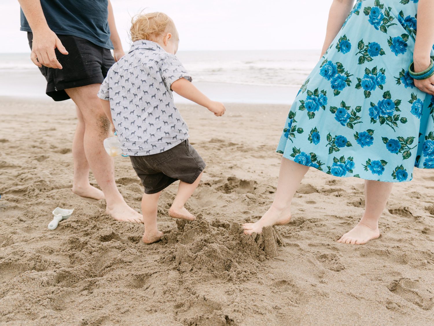 Family with toddler playing barefoot on sandy beach, child in floral shirt between parents in summer outfits.