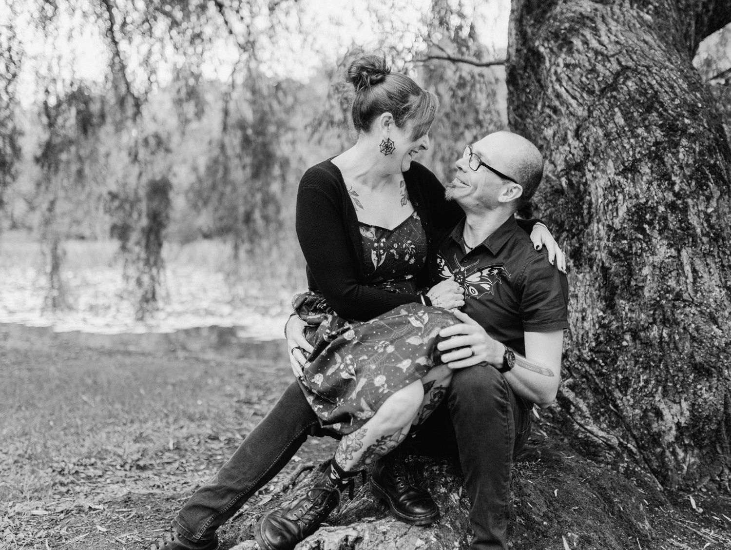 Black and white photo of a couple sitting against a tree, sharing a romantic moment outdoors near a pond.