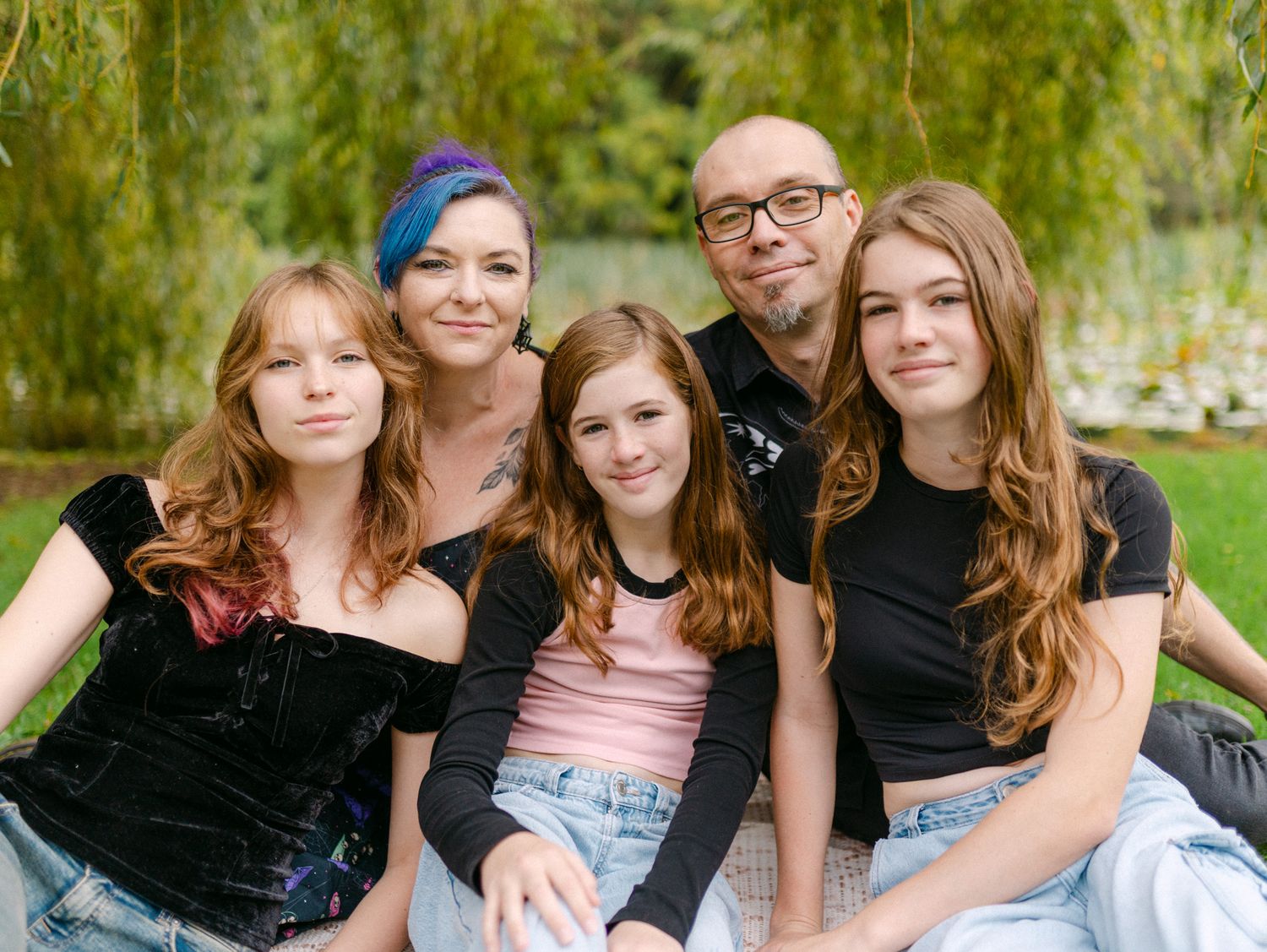 Family of five smiling outdoors near a weeping willow tree, posing together for a portrait photo.