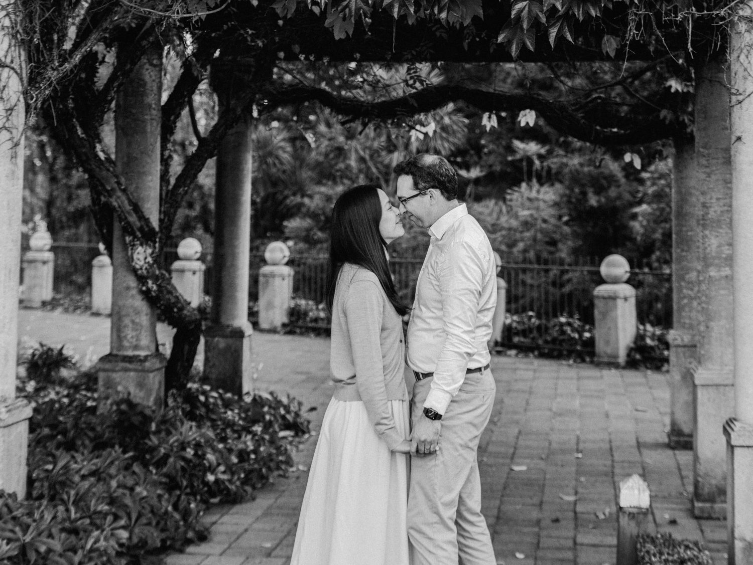 Black and white photo of a couple sharing a romantic moment under a garden pergola surrounded by lush greenery.