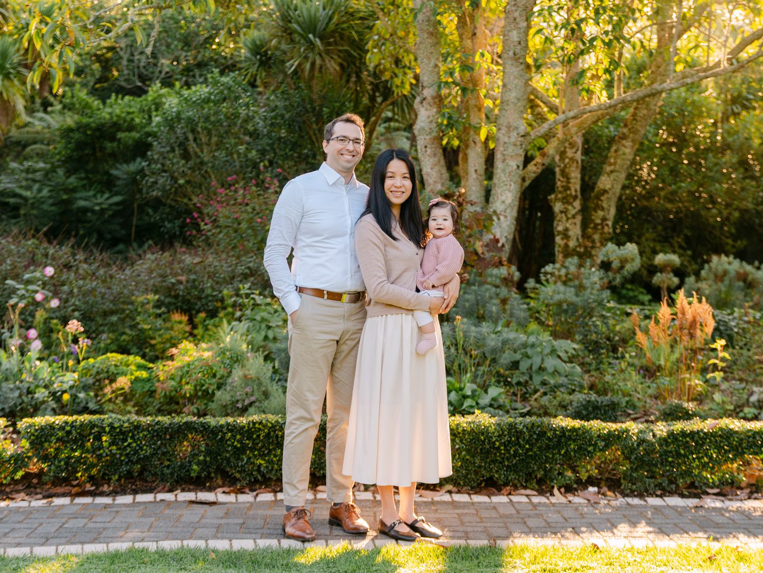 Family portrait of couple with baby in lush garden setting, wearing neutral tones on brick pathway.
